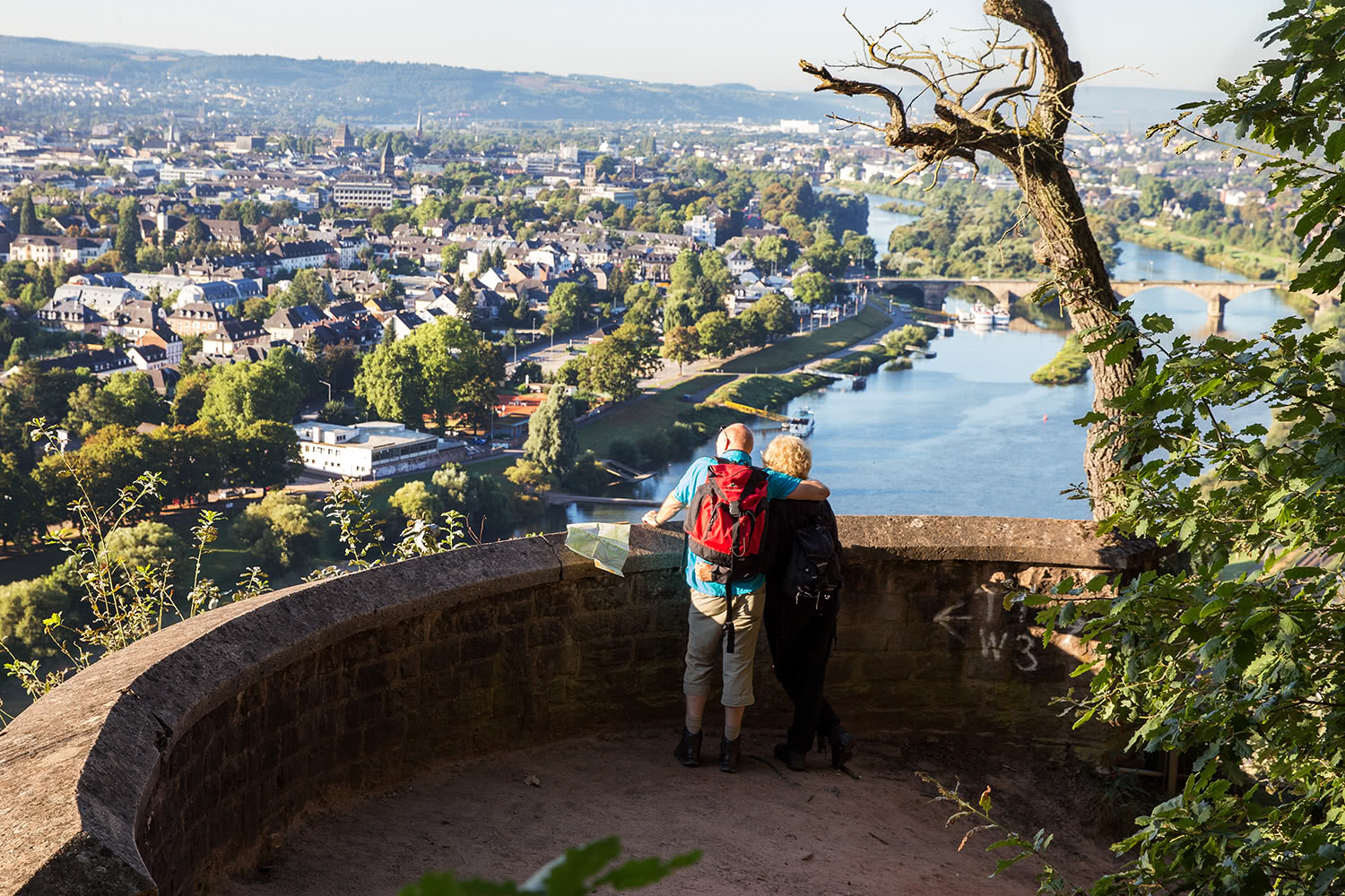 Bild von zwei Personen die an einer Mauer einer Aussiechtsplatform des Mosel Steig stehen und sich die Landschaft anschauen. Zu sehen ist die Mosel mit Blick auf Trier